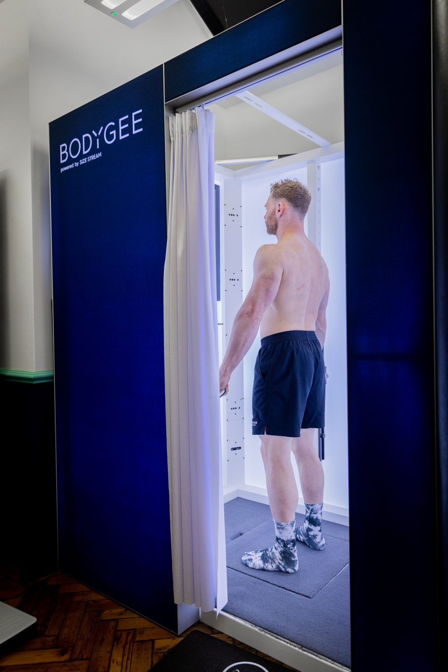 0K8A8738 The photo shows a white man having a body composition scan in a DEXA scanner. He is lying flat on his back on a raised table as the arm of the scanner passes over his body. There is a logo on the wall behind him that reads My Vital Metrics.