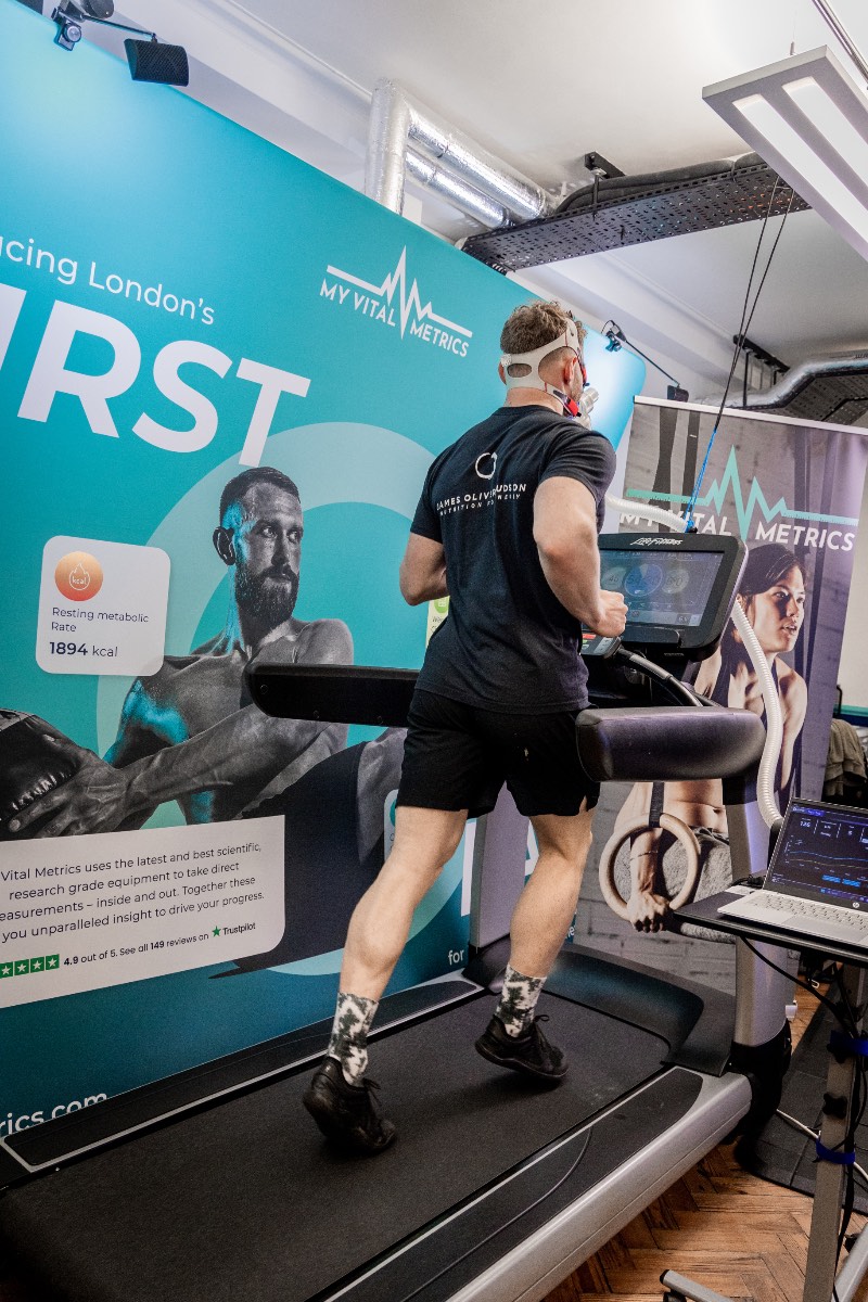 hudsonvo2 The photo shows a white man having a body composition scan in a DEXA scanner. He is lying flat on his back on a raised table as the arm of the scanner passes over his body. There is a logo on the wall behind him that reads My Vital Metrics.