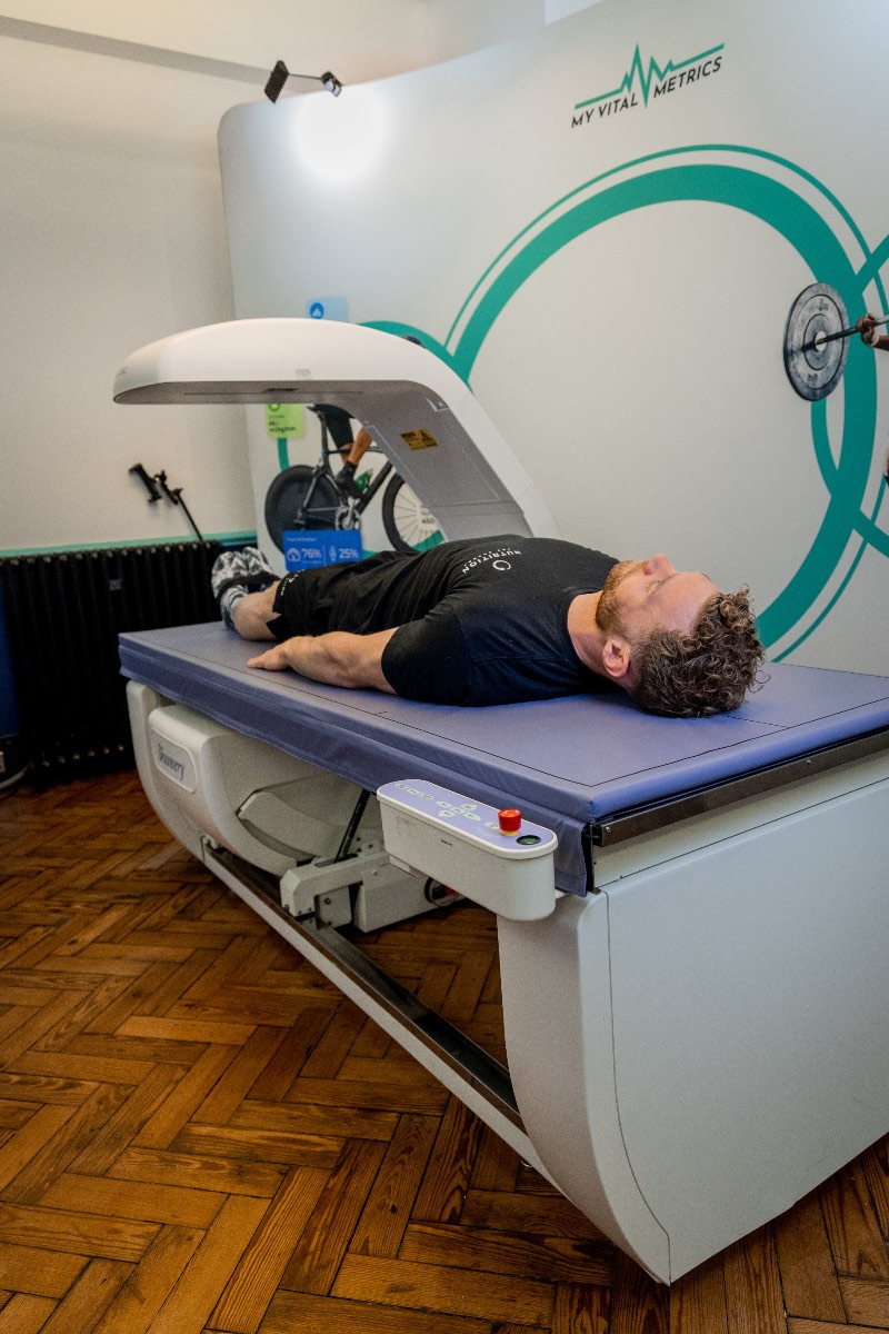 jamesH-dexa The photo shows a white man having a body composition scan in a DEXA scanner. He is lying flat on his back on a raised table as the arm of the scanner passes over his body. There is a logo on the wall behind him that reads My Vital Metrics.