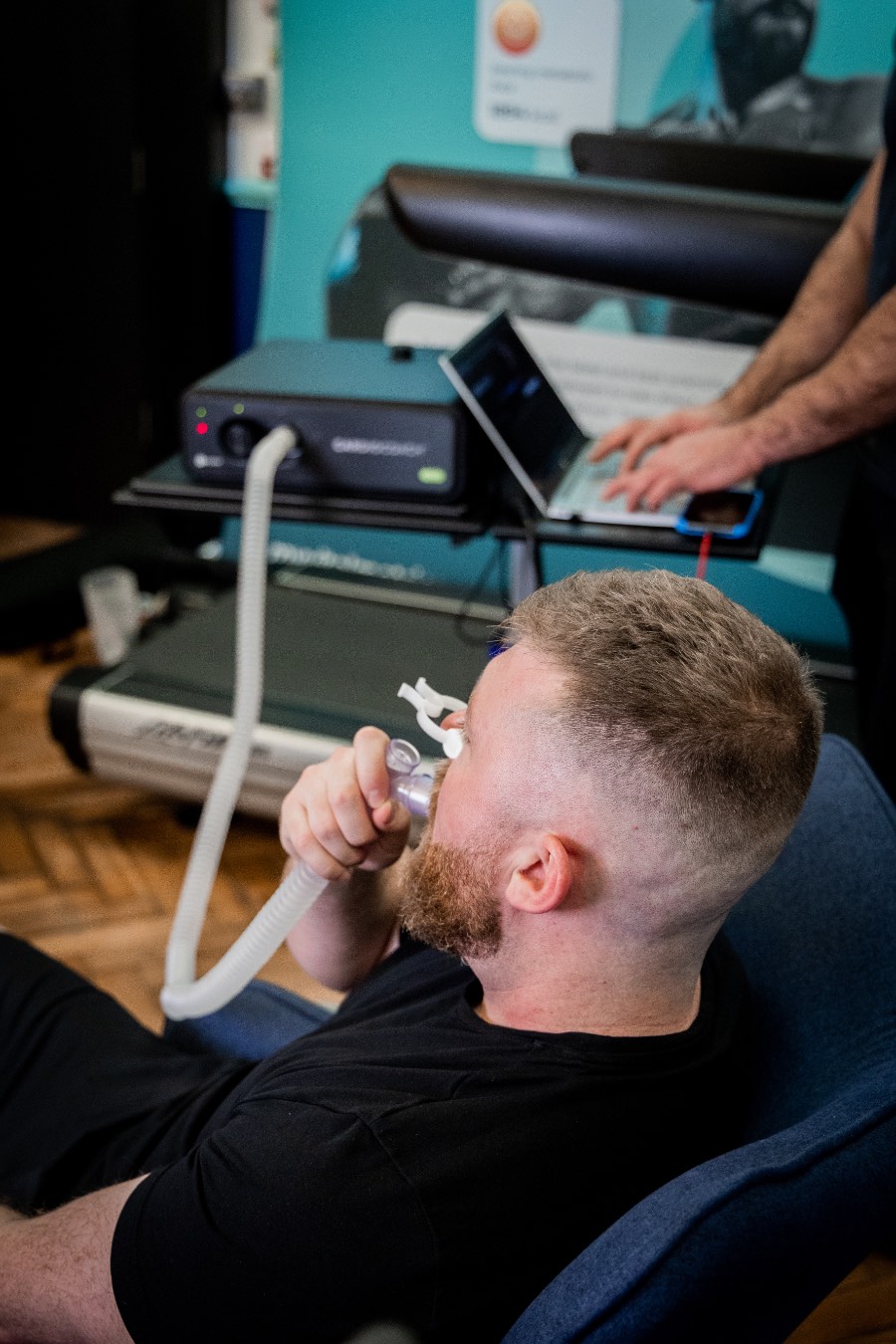 RMR in progress The photo shows a white man having a body composition scan in a DEXA scanner. He is lying flat on his back on a raised table as the arm of the scanner passes over his body. There is a logo on the wall behind him that reads My Vital Metrics.